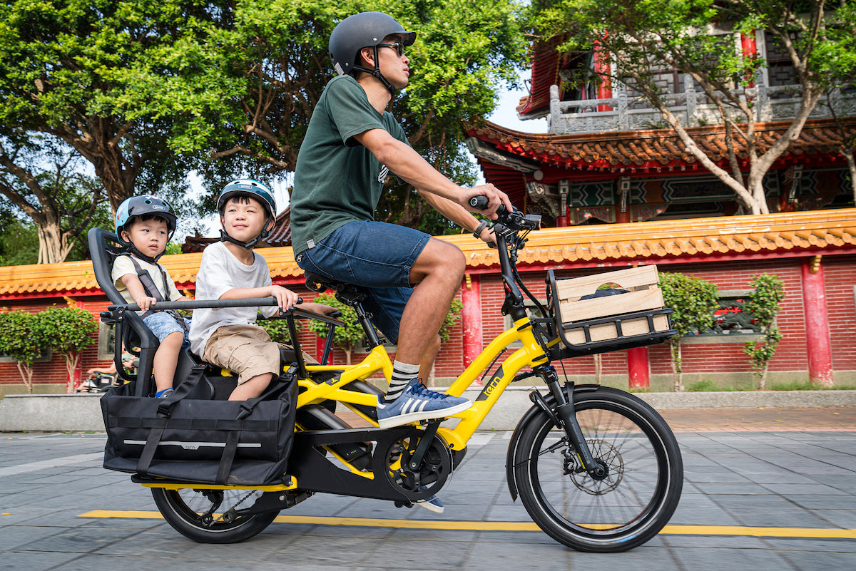 Man riding a yellow Tern electric bike with two children in a trailer, traditional building in the background